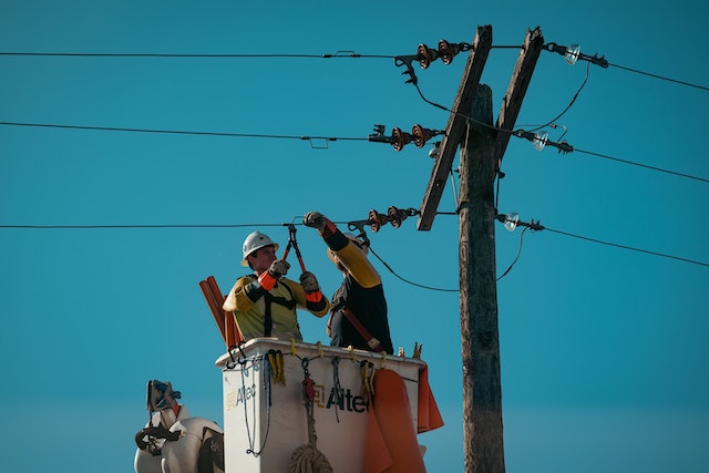 "Two utility workers in a bucket lift repairing power lines on a utility pole against a clear blue sky."