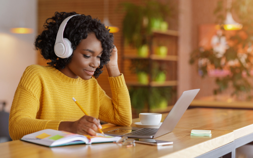 "A woman wearing headphones studies at a laptop while taking notes in a notebook, sitting at a wooden table in a cozy, plant-filled room."